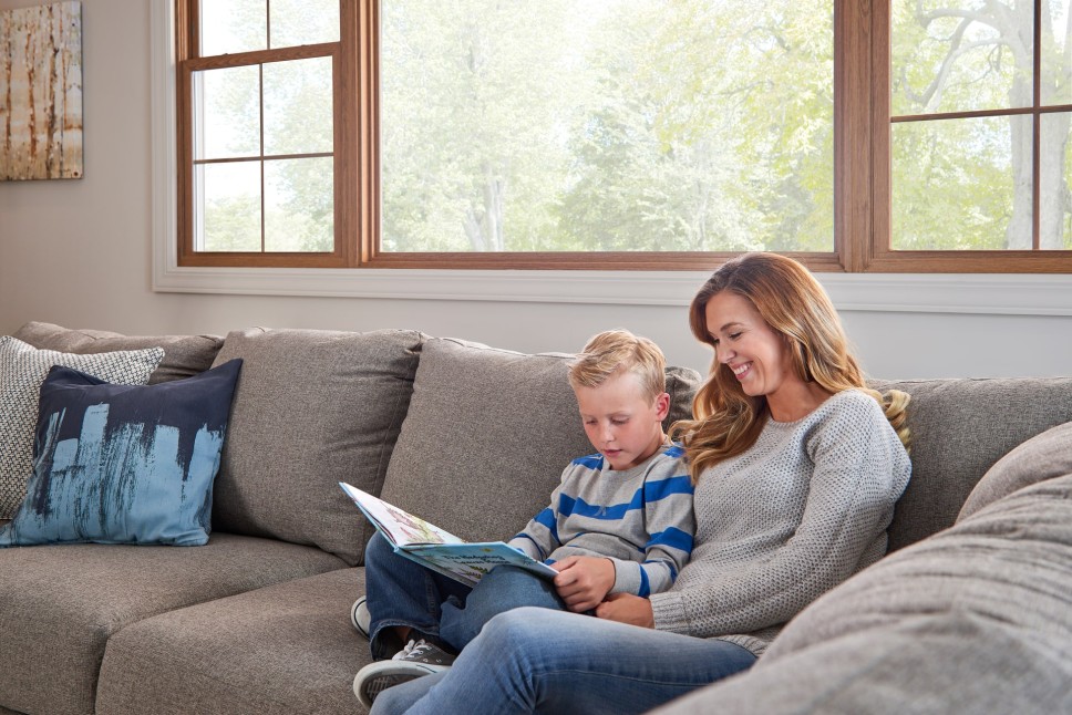 A woman and a child sitting on a couch in front of a large window inside a home